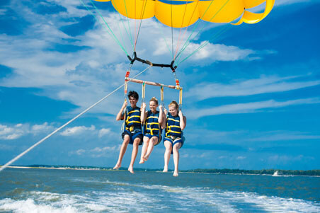 A full shot of a man and two women under a parasail wing wearing a blue life jacket against a blue sea and sky background.